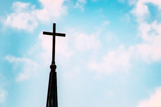 A church spire with a cross silhouetted against a bright blue sky with clouds.